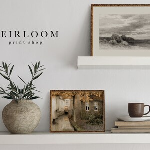 May include: A display of framed art prints and decorative items on a white shelf. The prints include a landscape sketch and a street scene. A ceramic vase holds olive branches. The text "HEIRLOOM print shop" is visible.