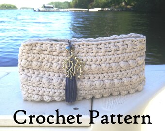 May include: A crocheted clutch bag in a cream color with a gold coral charm and a dark blue tassel. The bag is sitting on a white surface with a blurry background of a lake and trees.