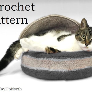 May include: A cat rests in a gray, beige, and dark gray cat bed. The bed has a rounded shape with a curved top. The cat is white and brown, with its tail extending out of the bed. The words "Crochet Pattern" are at the top.