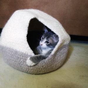 May include: A gray and white cat is curled up inside a gray and white knitted cat bed. The cat bed is shaped like a dome with a small opening.