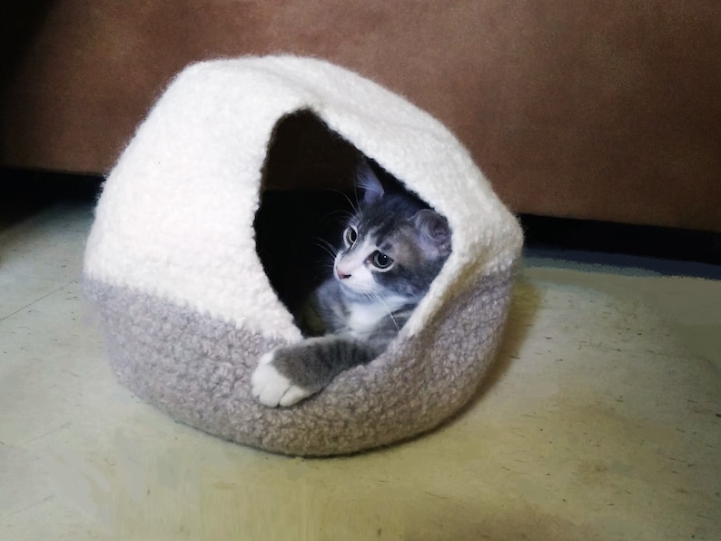May include: A gray and white cat is curled up inside a gray and white knitted cat bed. The cat bed is shaped like a dome with a small opening.