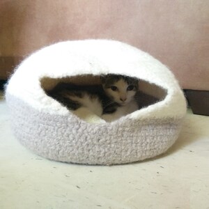 May include: A white and gray crocheted cat bed with a cat inside. The cat bed is shaped like a dome and has a small opening for the cat to enter.