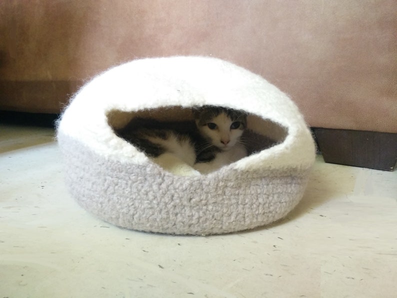 May include: A white and gray crocheted cat bed with a cat inside. The cat bed is shaped like a dome and has a small opening for the cat to enter.