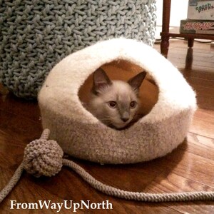 May include: A gray and white knitted cat bed with a gray rope toy. A gray and white cat is inside the bed looking out.