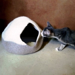 May include: A gray and white knitted cat bed shaped like a dome with a small opening. A gray and white cat is looking into the opening of the bed.
