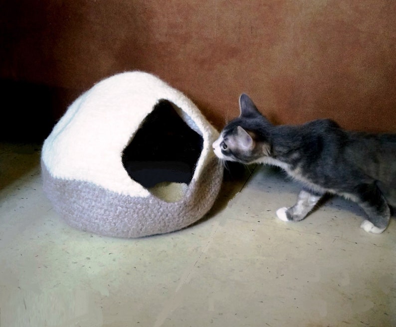 May include: A gray and white knitted cat bed shaped like a dome with a small opening. A gray and white cat is looking into the opening of the bed.