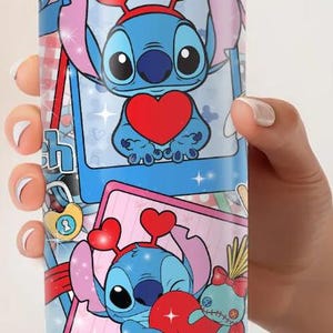 May include: A stainless steel tumbler featuring a cartoon character with a blue body, large eyes, and heart-shaped accents. The design includes the character holding a heart, set against a pink and blue background with the word "Love".