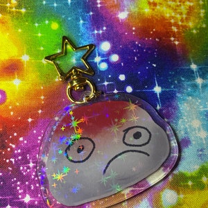 Puede incluir: Un llavero holográfico transparente con un personaje de dibujos animados. El personaje tiene una expresión triste, con ojos negros y una boca curvada. El llavero tiene un cierre dorado en forma de estrella.