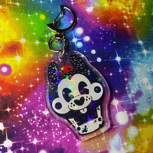 May include: A black and white cartoon character keychain with a red nose and sprinkles. The keychain is made of clear plastic with a holographic finish and has a silver metal ring.