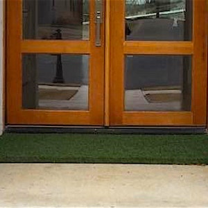 May include: A green artificial grass doormat placed in front of a wooden double door with glass panels.