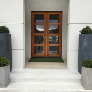 May include: A white modern home with a wooden double door entryway. Two large gray planters with green plants are flanking the door.
