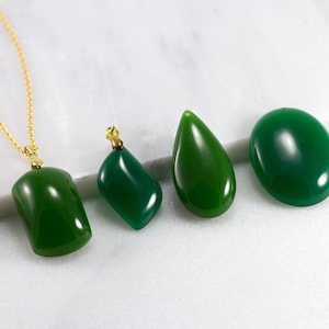 May include: Four green gemstone pendants with gold bail settings. The pendants are in various shapes, including oval, teardrop, and rectangular. The pendants are displayed on a white marble surface.