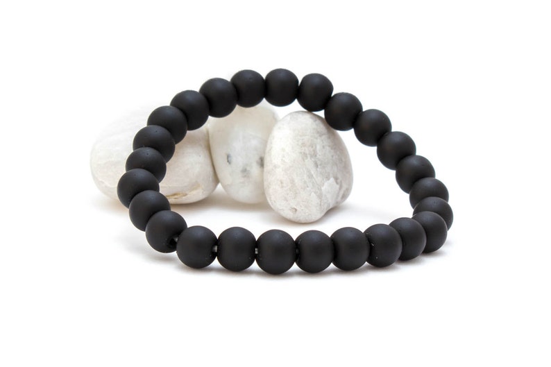May include: A black beaded bracelet with a matte finish. The bracelet is made of round beads and is lying on a white surface.