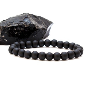 May include: A black beaded bracelet made of matte black beads. The bracelet is lying on a white surface next to a black rock.