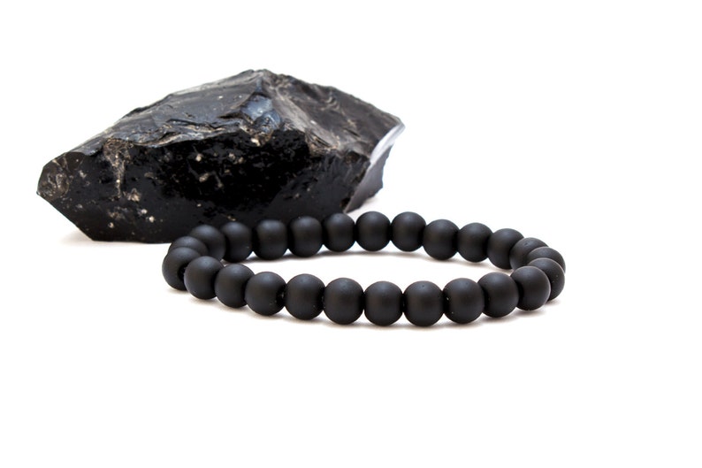 May include: A black beaded bracelet made of matte black beads. The bracelet is lying on a white surface next to a black rock.