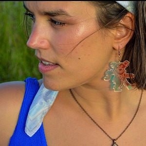 May include: A woman wearing a blue swimsuit, a white and blue patterned headband, and a necklace with a brown pendant. The earrings are clear with a dancing bear design in red and blue.