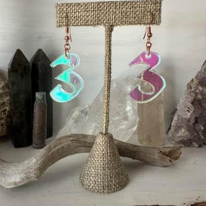 May include: A pair of iridescent earrings shaped like the number 3. The earrings are hanging from a brown fabric stand.