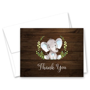 May include: A thank you card with a watercolor illustration of a gray elephant with blue polka dots sitting in a green wreath on a brown wood background. The text "Thank You" is written in cursive below the elephant.