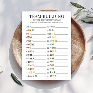 May include: A white printed game sheet titled "TEAM BUILDING OFFICE PICTIONARY GAME" with emoji clues. The sheet is on a wooden plate with olive branches. The game is designed to guess workplace phrases.