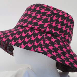 K&ouml;nnte beinhalten: Ein breitkrempiger Hut mit einem schwarz-pinkfarbenen Hahnentrittmuster. Die Unterseite des Krempe weist ein Paisley-Design in Rot-, Rosa- und Braunt&ouml;nen auf. Der Hut wird auf einem wei&szlig;en Schaufensterkopf pr&auml;sentiert.