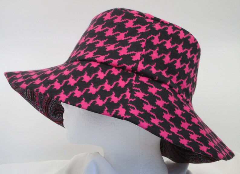 K&ouml;nnte beinhalten: Ein breitkrempiger Hut mit einem schwarz-pinkfarbenen Hahnentrittmuster. Die Unterseite des Krempe weist ein Paisley-Design in Rot-, Rosa- und Braunt&ouml;nen auf. Der Hut wird auf einem wei&szlig;en Schaufensterkopf pr&auml;sentiert.