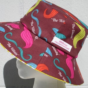 May include: A maroon bucket hat with a yellow brim and a colorful pattern of mustaches and text. The text includes "The Classic", "The Villain", "The Dandy", and "The Wandy".
