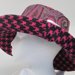 K&ouml;nnte beinhalten: Ein breitkrempiger Hut mit auff&auml;lligem Design. Der Rand weist ein schwarz-pinkfarbenes Hahnentrittmuster auf, w&auml;hrend die Krone einen Paisley-Druck in Rosa-, Rot- und Lilat&ouml;nen zeigt. Der Hut wird auf einem wei&szlig;en Schaufensterkopf pr&auml;sentiert.