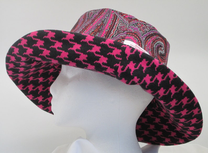 K&ouml;nnte beinhalten: Ein breitkrempiger Hut mit auff&auml;lligem Design. Der Rand weist ein schwarz-pinkfarbenes Hahnentrittmuster auf, w&auml;hrend die Krone einen Paisley-Druck in Rosa-, Rot- und Lilat&ouml;nen zeigt. Der Hut wird auf einem wei&szlig;en Schaufensterkopf pr&auml;sentiert.