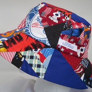 Op de afbeelding: Een blauwe en rode patchwork-emmerhoed met verschillende patronen, waaronder sterren, strepen en een baseball-logo. De hoed heeft een label met de tekst "SR Designs" en contactgegevens.