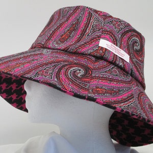 K&ouml;nnte beinhalten: Ein breitkrempiger Hut mit einem Paisley-Muster in Rosa-, Lila- und Schwarzt&ouml;nen. Die Unterseite der Krempe hat ein schwarz-rosa Hahnentrittmuster. Der Hut hat ein Etikett mit Text.