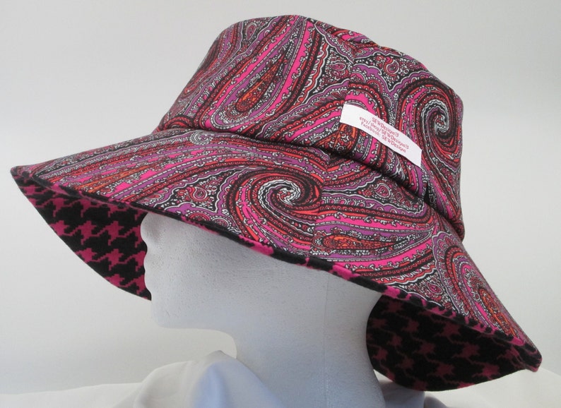 K&ouml;nnte beinhalten: Ein breitkrempiger Hut mit einem Paisley-Muster in Rosa-, Lila- und Schwarzt&ouml;nen. Die Unterseite der Krempe hat ein schwarz-rosa Hahnentrittmuster. Der Hut hat ein Etikett mit Text.