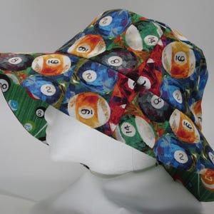 May include: A wide-brimmed hat with a colorful pattern of billiard balls. The balls are painted in various shades of red, yellow, blue, green, and black. Each ball has a white number on it.