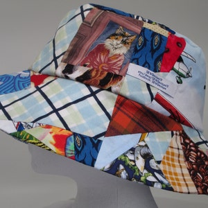 May include: A wide-brimmed sun hat made from patchwork fabric. The hat is blue, red, green, and white with a variety of patterns, including plaid, floral, and geometric designs. The hat has a label that reads "SF Designs" and includes a website and Facebook page.