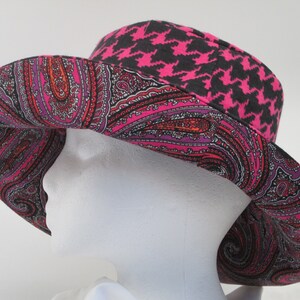 K&ouml;nnte beinhalten: Ein breitkrempiger Hut mit einem lebendigen Paisley-Muster in Pink, Rot und Lila. Der Hut hat ein schwarzes und pinkfarbenes Hahnentrittmuster auf der Krone. Der Hut wird auf einem wei&szlig;en Schaufensterkopf pr&auml;sentiert.