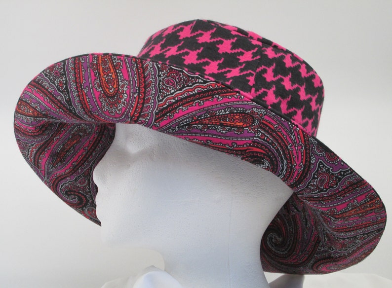 K&ouml;nnte beinhalten: Ein breitkrempiger Hut mit einem lebendigen Paisley-Muster in Pink, Rot und Lila. Der Hut hat ein schwarzes und pinkfarbenes Hahnentrittmuster auf der Krone. Der Hut wird auf einem wei&szlig;en Schaufensterkopf pr&auml;sentiert.