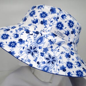 May include: White sun hat with a blue floral pattern and a blue trim. The hat features a wide brim and a tag with text. The hat is designed to provide sun protection and is suitable for outdoor activities.