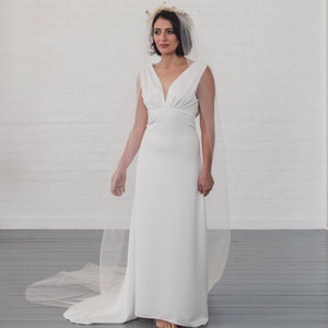 Puede incluir: Un vestido de novia blanco con cuello en V y falda larga y fluida. El vestido se combina con un velo transparente y una corona floral. El vestido es sencillo y elegante, para una novia.