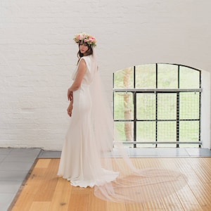 Puede incluir: Una novia con un vestido de novia blanco, un velo largo y una corona de flores. La novia está de pie frente a una ventana con vista a los árboles.