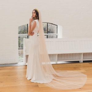 Pode incluir: Uma noiva usando um vestido de noiva branco com um véu longo e esvoaçante. O véu é de cor rosa claro e tem uma textura delicada e transparente. A noiva está em pé em um quarto branco com um piso de madeira.
