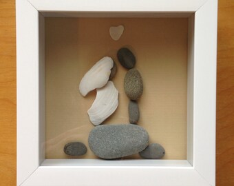 以下が含まれることがあります： 石と貝殻でできた額装されたアート作品。カップルが抱きしめ合い、その上に白いハートが描かれている。作品は薄い茶色の木製の表面に飾られている。