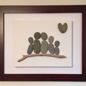 May include: A framed artwork depicting a family of five represented by smooth gray stones sitting on a driftwood branch. A broken heart made of stones is in the upper right corner of the artwork.