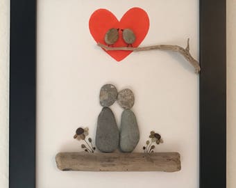 May include: A framed artwork featuring a couple made of grey stones sitting on a piece of driftwood. Two small birds are perched on a branch above them, with a red heart painted in the background.