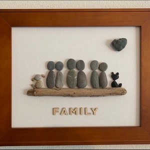 May include: A framed artwork made with stones depicting a family of six people and a dog sitting on a driftwood log. The word "FAMILY" is spelled out in wooden letters below the log. A heart-shaped stone is in the upper right corner of the image.