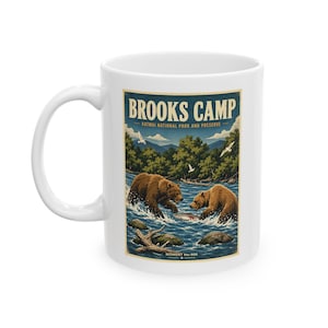 Op de afbeelding: Een witte keramische mok met een grafische illustratie van twee bruine beren die in een rivier vissen. De tekst luidt BROOKS CAMP, KATMAI NATIONAL PARK AND PRESERVE en MOMENT No. 006. De afbeelding toont groene bomen, blauwe bergen en witte vogels.
