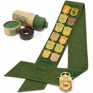 May include: A green sash with wooden badges featuring national park names, including Zion and Arches. The sash is accompanied by a roll of green fabric, a roll of brown ribbon, and a wooden Junior Park Ranger badge. The words "DISCOVER NATIONAL" are printed on the packaging.