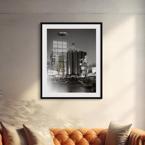 May include: A black and white photograph of a city skyline with a flying saucer in the sky. The image is framed in a black frame and hanging on a wall.