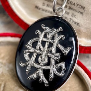 May include: Oval black pendant with a silver-colored, intertwined design. The pendant hangs from a silver chain. The pendant is displayed in a red-lined box with text that is partially visible.