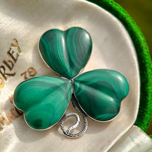May include: A green malachite shamrock brooch with a silver-tone metal setting. The three-leaf clover design is set against a cream-colored satin background inside a green velvet-lined box. The brooch is a unique piece of jewelry.