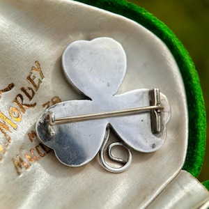 May include: A silver-toned shamrock brooch, featuring three heart-shaped leaves and a curled stem. The brooch is set against a cream-colored satin lining within a green velvet-lined box. The box has gold lettering.
