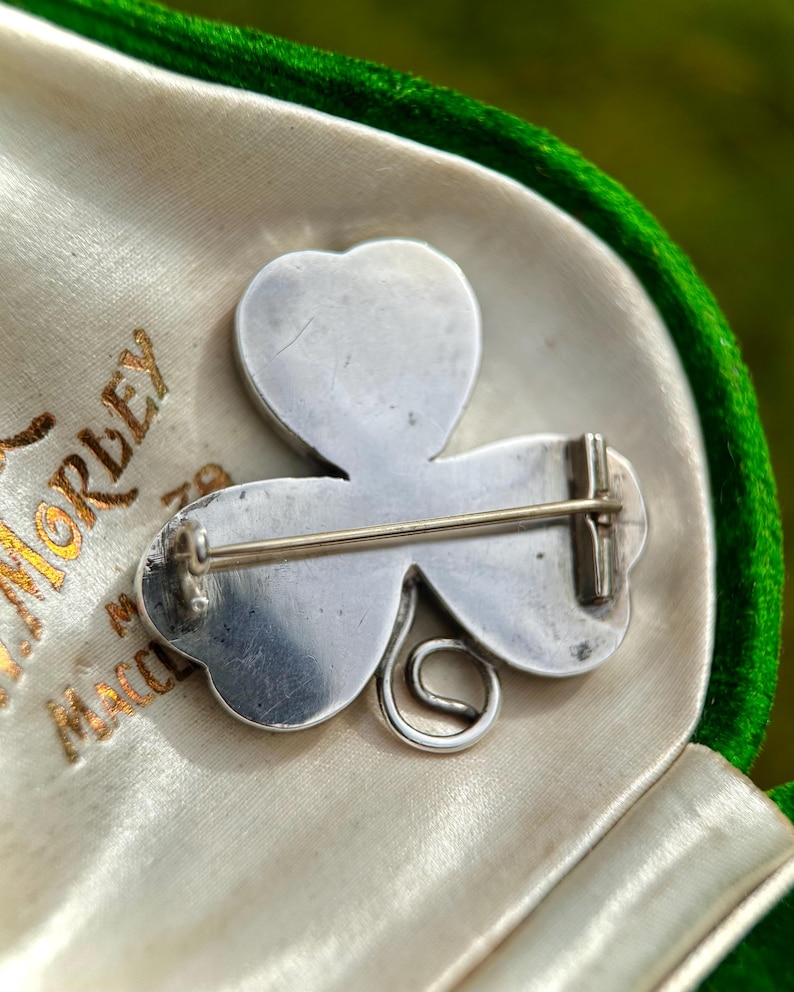 May include: A silver-toned shamrock brooch, featuring three heart-shaped leaves and a curled stem. The brooch is set against a cream-colored satin lining within a green velvet-lined box. The box has gold lettering.